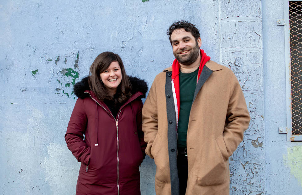 Ric and Morgan from the Adhoc Presents team stand smiling against a light blue wall. 